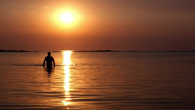 The silhouette of an athletic man comes out of the water and goes in the direction of the camera. The solar disk rises above the sea