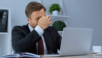 Naklejka premium exhausted businessman rubbing eyes while suffering from migraine near laptop