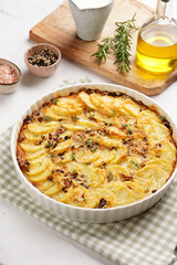Slices potato casserole with red onions, fresh thyme and rosemary on green checkered kitchen towel , wooden board with flavoring ingredients: pepper, salt, olive oil