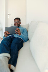 Shot of a handsome young man using his digital tablet while sitting on a sofa at home. Working with mobile gadget. Young African-American man using digital tablet, sitting on sofa at home, copy space