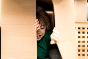 Boy hiding in inside a huge cardboard box. He is playing and peeking through a hole in box. Kid is happy about moving into a new home.