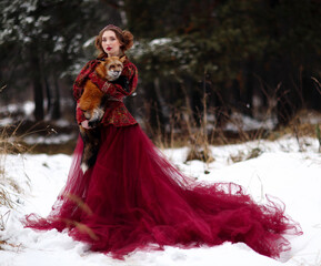 Beautiful young woman in red traditional dress with red fox on hands in winter forest