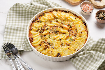 Slices potato casserole with red onions, fresh thyme and rosemary on green checkered kitchen towel , wooden board with flavoring ingredients: pepper, salt, olive oil