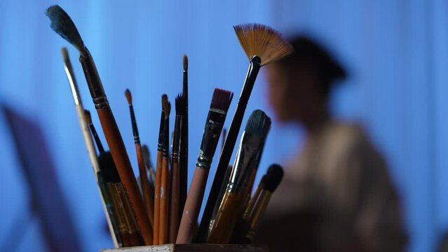 Focus on artist's art tools in dark studio workshop. Brushes, paintbrush, palette knife close up. In the background, the blurred outlines of an artist painting a picture on canvas. Slow motion.