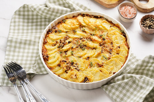 Slices potato casserole with red onions, fresh thyme and rosemary on green checkered kitchen towel , wooden board with flavoring ingredients: pepper, salt, olive oil