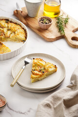 Slice of potato casserole with red onions, fresh thyme and rosemary on green checkered kitchen towel , wooden board with flavoring ingredients: pepper, salt, olive oil