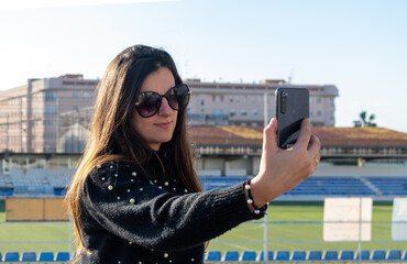 mujer  caucásica con pelo liso castaño haciendo videollamada dentro de un estadio de futbol, con gafas de sol y camiseta negra.