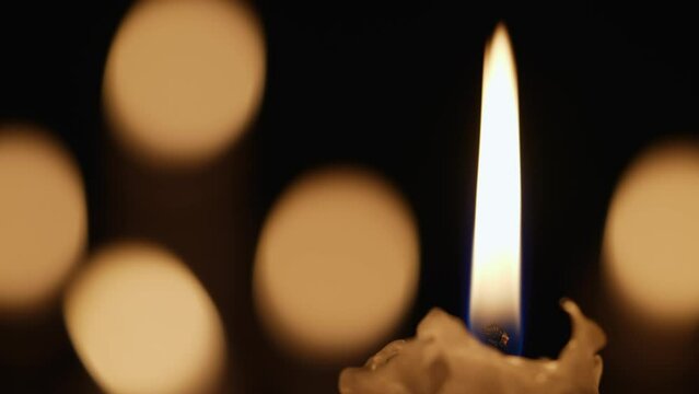 Wax burning candle flickers in dark on a black background with blurred candlelight bokeh effect. Background or illustration of remembrance, mourning or celebration. Blaze candles in church close up.