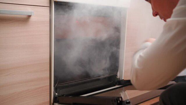 Man Opens an Oven Full of Smoke in the Kitchen. Male Bachelor Ruined Dinner with Burnt Food. Bad Cook. 
