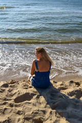 Fototapeta na wymiar Young woman sits on the sand on the beach near the sea and looks into the distance