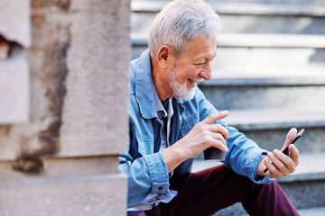 A happy senior student is sitting on the stairs with a phone and coffee in his hands and takes a...