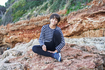 The boy looking at camera is sitting on a rock by the sea. - Powered by Adobe