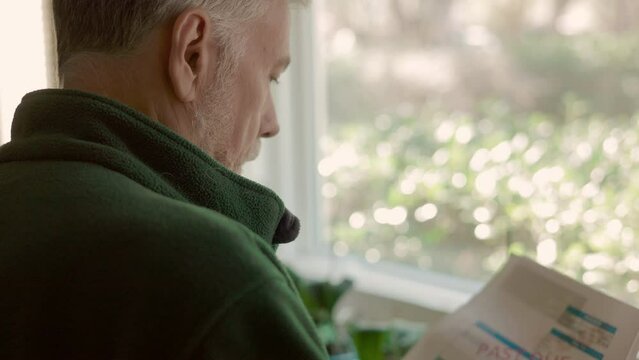 View over the shoulder of a solemn senior citizen standing by a window looking at some past due notices.
