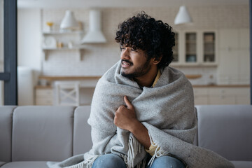 Sick unhappy Indian man covered with plaid blanket sitting in cold apartment looking away. Illness, quarantine, self isolation concept - obrazy, fototapety, plakaty
