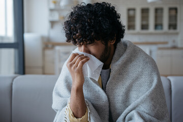 Sick Indian man with runny nose holding paper napkins near face sitting at home. Flu, virus,...