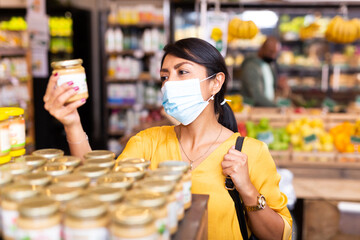Woman in protective mask reads description of canned food at a grocery supermarket