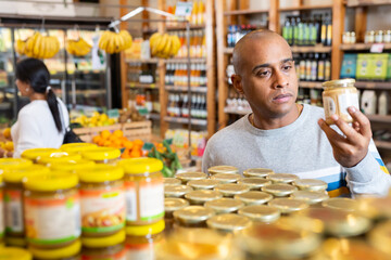 Interested Latin American man reading product label on jar while choosing groceries in supermarket