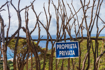 A wooden fence built of dry branches against the background of a green hill and blue sea. On the fence a blue sign with the Italian text "proprieta privata" which means "private property"