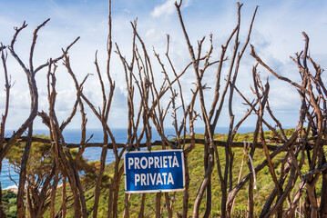 A wooden fence built of dry branches against the background of a green hill and blue sea. On the fence a blue sign with the Italian text "proprieta privata" which means "private property"