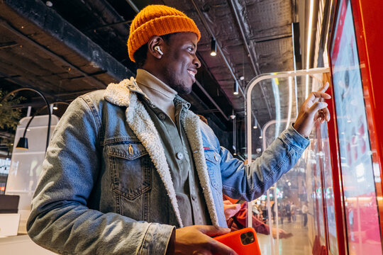 Smiling African-American man in warm denim jacket with wireless earphones uses self-service kiosk to order snack in cafe