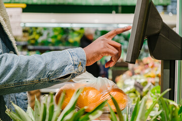 Stylish African-American guy in warm jacket with disposable mask weighs fresh oranges with digital scale in light supermarket