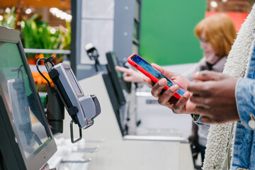 Black male person in warm denim jacket uses smartphone to pay for purchase at self-checkout point...
