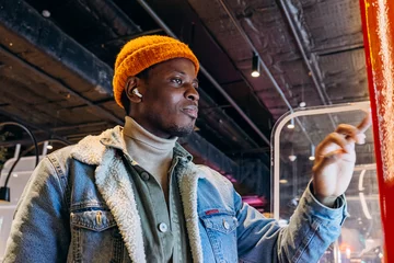 Deurstickers Positive African-American guy in orange knitted hat with wireless earphones orders food through self-service kiosk in cafe © wifesun