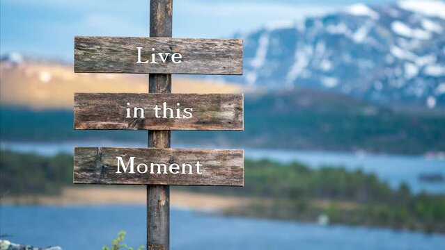 live in this moment text on wooden signpost outdoors in landscape scenery during blue hour. Sunset light, lake and snow capped mountains in the back.