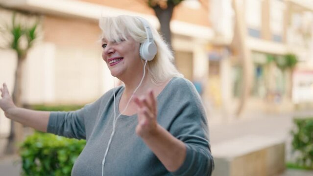 Middle age blonde woman listening to music using smartphone and dancing at street