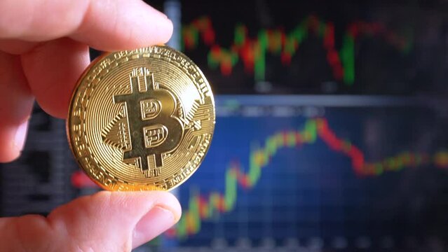 Male finger holding golden Bitcoin in light and stock index curves in background,close up