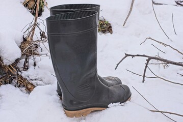two green rubber boots stand on a winter street in white snow and dry grass and branches
