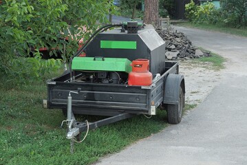 a black car trailer with a mobile generator and a red gas bottle stands on the street on gray asphalt and in green grass