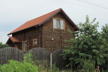 facade of a brown wooden house with windows under a tiled roof behind a fence on a rural street in green vegetation
