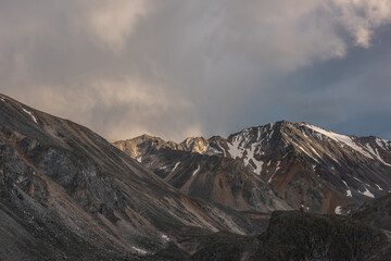 Fototapeta premium Dramatic landscape with high mountain range with sunlit golden sharp rocky top under clouds of sunset color in gloomy sky. Dark atmospheric scenery with large mountains with snow at changeable weather