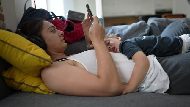Mother lying on sofa using cellphone with child asleep on her belly
