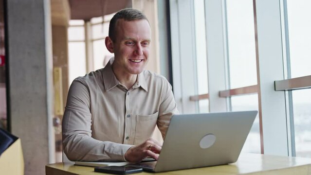 Smiling young man working at his laptop. Male employee says something and looks into window. Blurred backdrop.