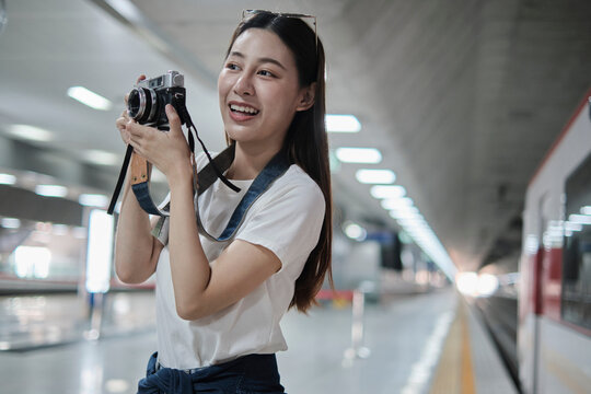 One young beautiful Asian female tourist, photographer taking snapshot photos with film camera, smile and enjoyment at train station platform, happy travel lifestyle by subway transport vacation trip.