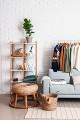 Shelf unit, sofa and rack with sweaters near white brick wall in living room interior