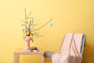 Vase with tree branches, Easter eggs on table and armchair near color wall
