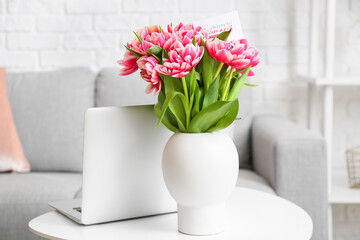 Modern laptop, vase with bouquet of tulips and greeting card on table. International Women's Day celebration