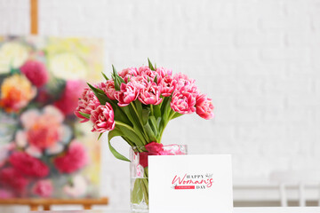 Vase with bouquet of beautiful tulips and greeting card with text HAPPY WOMAN'S DAY on table in light room
