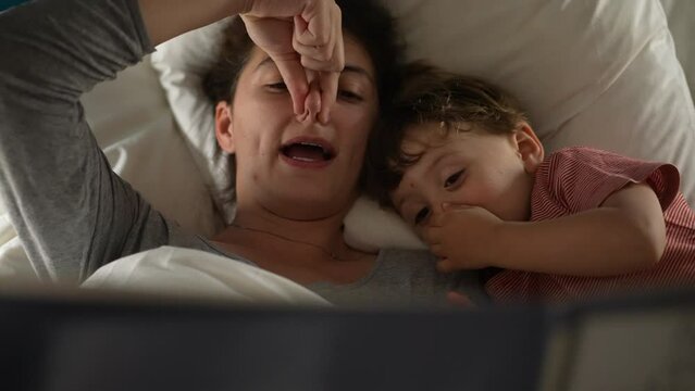 Playful mother telling a bedtime story to child son lying in bed imitating story