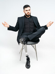 Disabled young man with prosthetic leg sitting on a chair in studio over white background,...