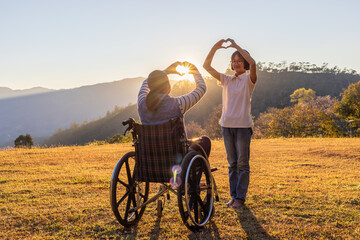 Young girl sitting in wheelchair and care helper playing on mountain meadow park in the sunset.