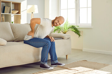 Unhealthy young man sit on couch at home struggle with backache. Pain in lower back. Unwell sick...