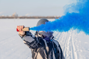 A male teenager in winter in an open space holds a smoke bomb with blue smoke in his hands. The concept of freedom and rebellion.