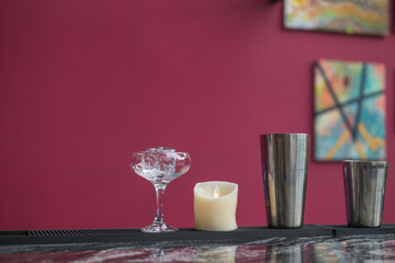 cooling the glass with ice before making a cocktail. Close up photo of dishes on the bar desk. Glass for cocktail, shakers, candle, pink background.