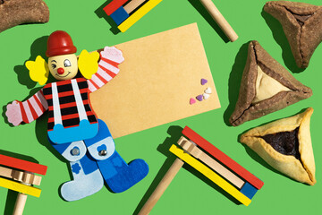 Decorative clown, blank notebook paper, Hamantaschen (traditional cookies for Jewish holiday Purim), wooden noisemaker on green background.