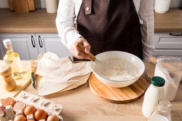 a woman's hand stirs the flour with a whisk in a bowl with the ingredients for the dough. butter, eggs, milk, a can of flour on the kitchen table.