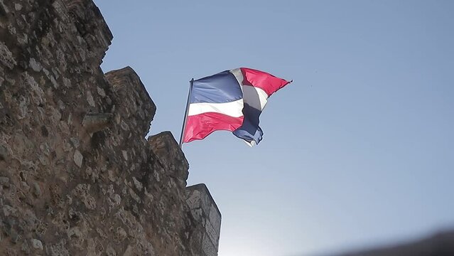 Captura a c&aacute;mara lenta de la bandera de la rep&uacute;blica dominicana ondeando en lo alto con un sol radiante detr&aacute;s y un cielo azul claro.